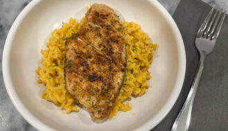 Close-up of creamy macaroni and cheese topped with sliced blackened chicken breast, garnished with parsley and served in a white bowl.