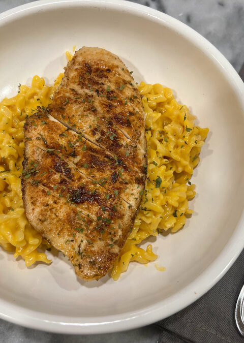 Close-up of creamy macaroni and cheese topped with sliced blackened chicken breast, garnished with parsley and served in a white bowl.