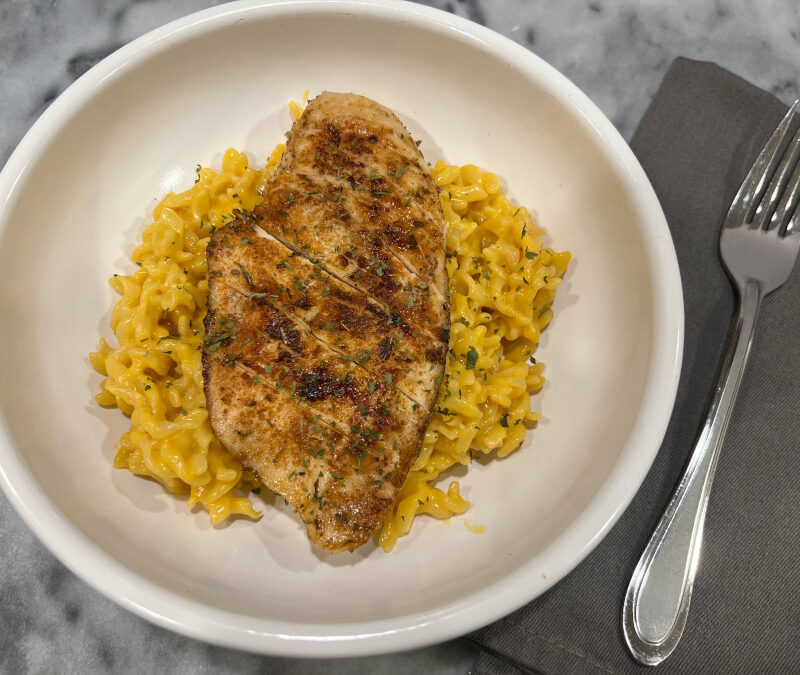 Close-up of creamy macaroni and cheese topped with sliced blackened chicken breast, garnished with parsley and served in a white bowl.