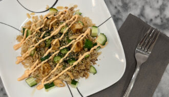 Vegetarian, plant-based sushi bowl with brown rice, diced cucumber, plant-based chicken, fried onions, and a drizzle of spicy mayo on a white plate.