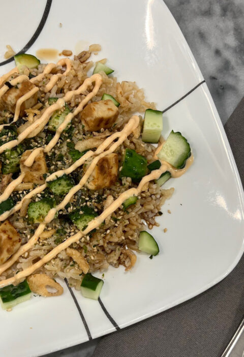 Vegetarian, plant-based sushi bowl with brown rice, diced cucumber, plant-based chicken, fried onions, and a drizzle of spicy mayo on a white plate.
