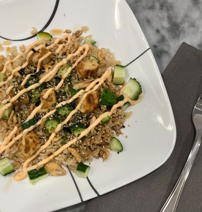 Vegetarian, plant-based sushi bowl with brown rice, diced cucumber, plant-based chicken, fried onions, and a drizzle of spicy mayo on a white plate.