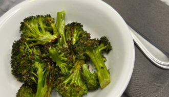 roasted broccoli in white bowl next to fork on gray napkin on gray and white marble background