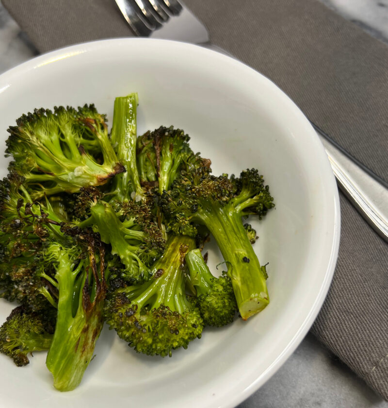 roasted broccoli in white bowl next to fork on gray napkin on gray and white marble background