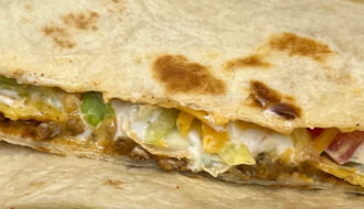 Vegetarian Crunchwrap Supreme cut in half, showing layers of plant-based meat, nacho cheese, tostada, lettuce, tomato, and sour cream inside a grilled tortilla wrap on a white plate