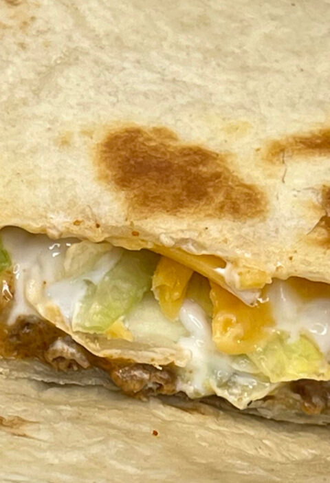 Vegetarian Crunchwrap Supreme cut in half, showing layers of plant-based meat, nacho cheese, tostada, lettuce, tomato, and sour cream inside a grilled tortilla wrap on a white plate
