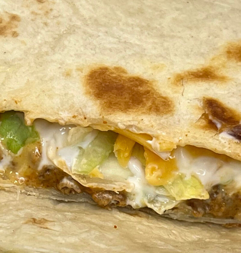 Vegetarian Crunchwrap Supreme cut in half, showing layers of plant-based meat, nacho cheese, tostada, lettuce, tomato, and sour cream inside a grilled tortilla wrap on a white plate