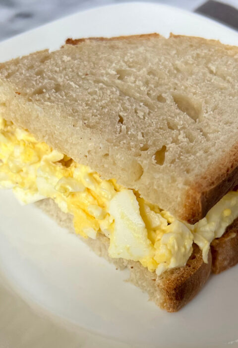 Close-up of a creamy egg salad sandwich on toasted bread, served on a white plate